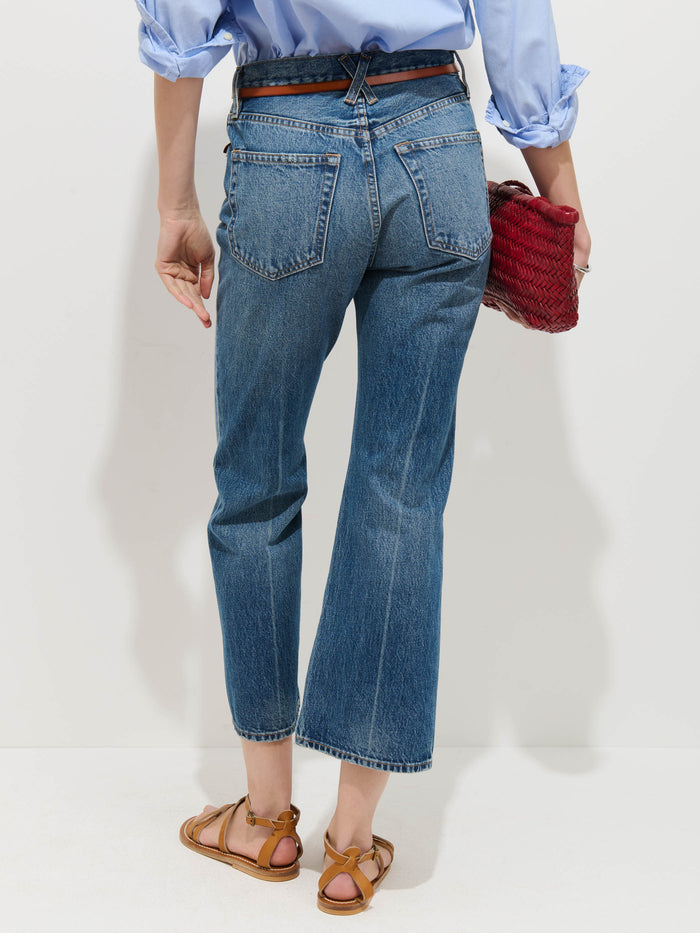 A person, shown from the back against a plain white background, wears Bessette Mid Rise Boot Cut Jeans, a light blue rolled-sleeve shirt, brown sandals, and holds a red woven clutch.