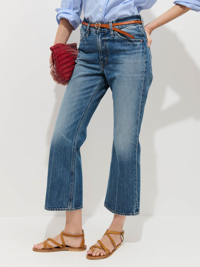 A person wearing Bessette Mid Rise Boot Cut Jeans with a light blue cotton shirt, tan strappy sandals, a brown belt, and holding a red textured clutch against a plain white background.