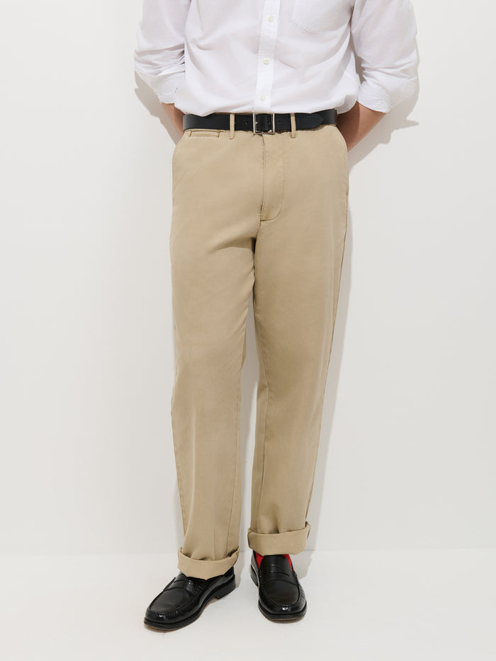 A person wearing a white button-down shirt, Flat Front Chino in Vintage Wash in beige with cuffs, a black belt, and black dress shoes stands against a plain white background.