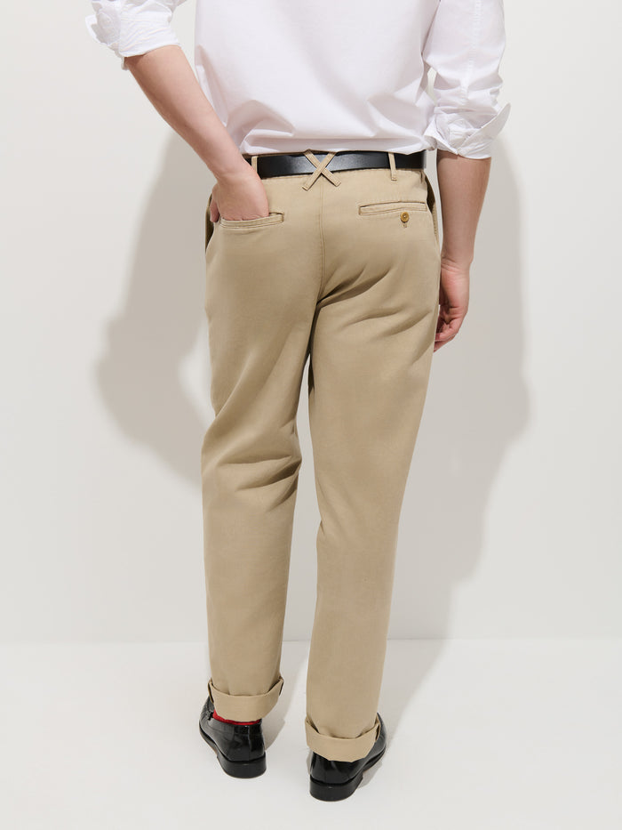 A person wearing the Flat Front Chino in Vintage Wash, paired with a black belt, black shoes, and a white shirt with rolled sleeves. Only the lower body is visible as they stand facing a white wall with one hand in a back pocket.