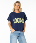 A woman with blonde hair wears the Roxy Free Time Top, a navy blue cozy cotton t-shirt featuring four yellow hibiscus flowers, paired with light blue ripped jeans. She stands with her hands in her pockets against a plain white background.