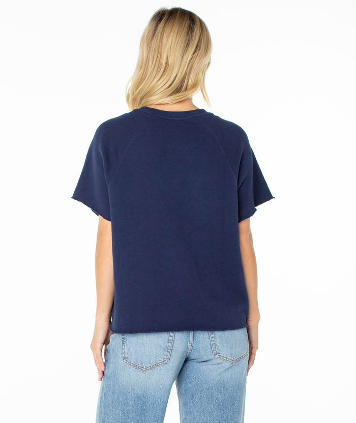 A person with long blonde hair, wearing the Roxy Free Time Top in navy with a relaxed silhouette and light blue jeans, stands facing away from the camera against a plain white background.