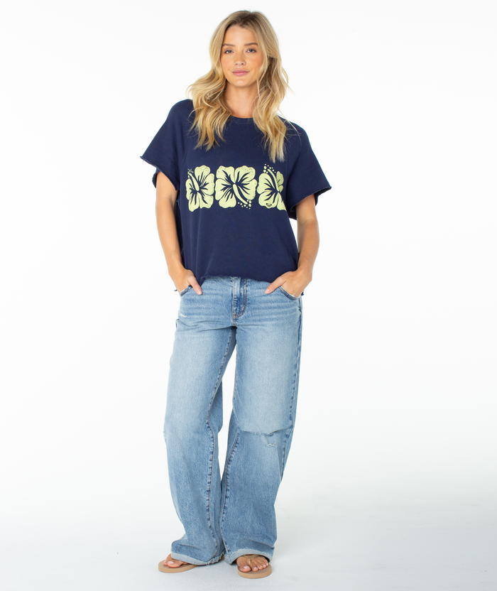 A woman wearing the Roxy Free Time Top, a navy blue cozy cotton t-shirt with yellow hibiscus flowers, paired with light blue jeans and sandals, stands against a white background with her hands in her pockets, looking at the camera.