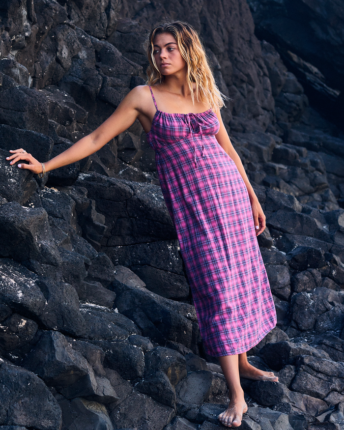 A woman wearing the Roxy Late Night Midi Dress, featuring a pink plaid pattern and a ruched drawstring neckline, stands barefoot on dark, rocky terrain with one hand resting against the rocks and her gaze turned to the side—an effortlessly elegant look ideal for relaxed beachside occasions.