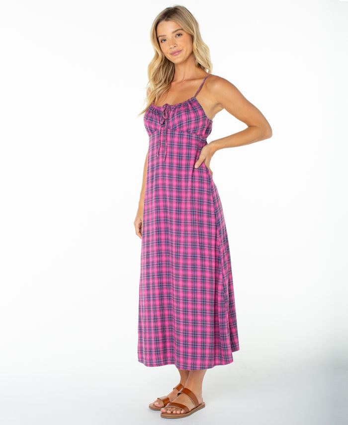A woman stands in the Roxy Late Night Midi Dress, featuring a pink and blue plaid pattern, spaghetti straps, and a ruched drawstring neckline. Paired with brown sandals against a white background, this sleeveless dress is perfect for any beachside occasion.