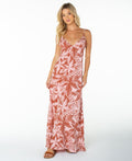 A woman wearing the Roxy Lost Paradise Dress in Chutney Bella Block—a long, sleeveless dress with spaghetti straps and a red and white floral pattern—paired with sandals, stands against a plain white background.