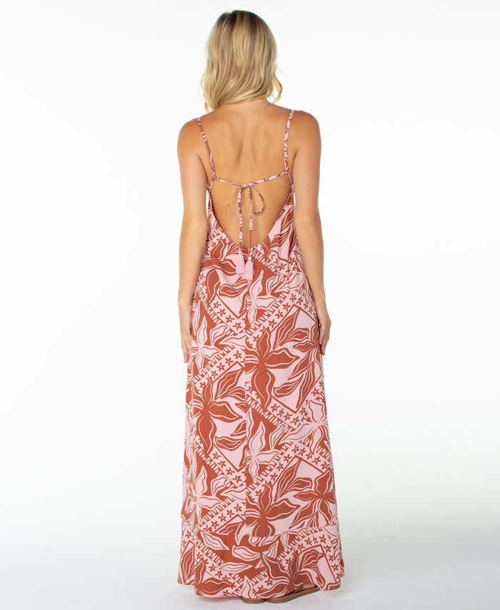 A woman with blonde hair models the Roxy Lost Paradise Dress, a pink and rust floral-patterned maxi dress featuring thin straps and an open back. She stands against a white background, facing away from the camera.