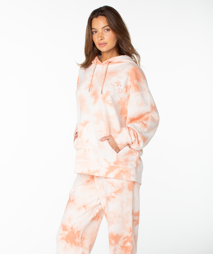 A woman stands with her hands in her pockets, wearing the Roxy Maile Tie Dye Hoodie—a relaxed, peach and white cotton sweatshirt—paired with matching sweatpants against a plain white background, embodying effortless loungewear style.