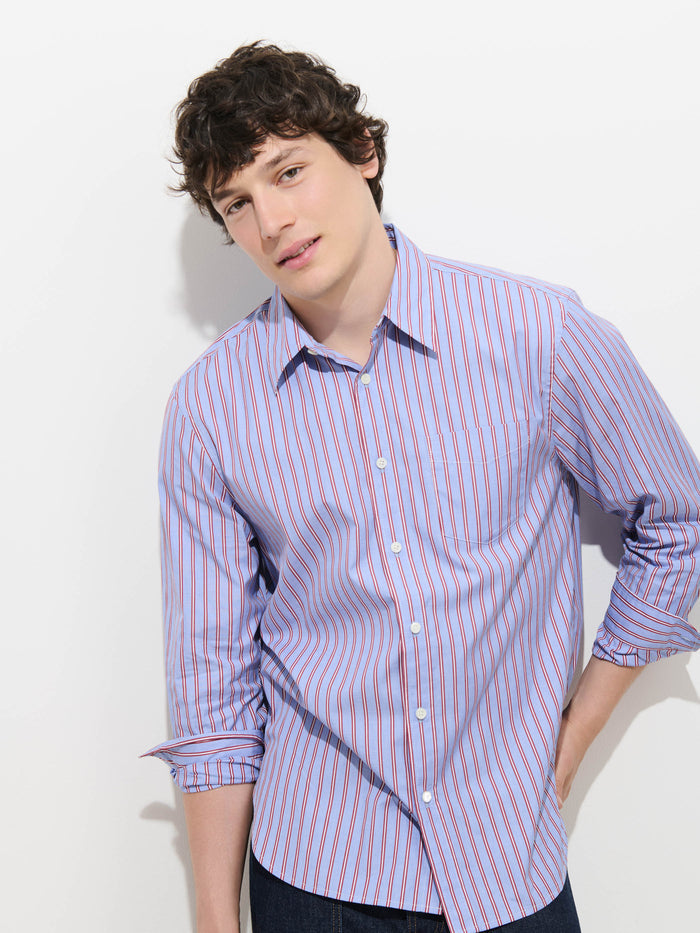 A young man with wavy brown hair is wearing the Mill Point Collar Striped Shirt in Cotton Poplin—a light blue, 100% cotton shirt with red stripes and a point collar—with the sleeves rolled up, standing against a white background with one hand in his pocket.