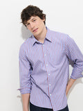 A young man with wavy brown hair is wearing the Mill Point Collar Striped Shirt in Cotton Poplin—a light blue, 100% cotton shirt with red stripes and a point collar—with the sleeves rolled up, standing against a white background with one hand in his pocket.