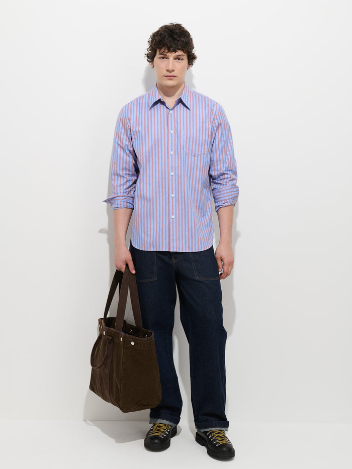 A person with short curly hair wears the Mill Point Collar Striped Shirt in Cotton Poplin—garment-dyed for a unique finish and crafted from 100% cotton—in blue and purple stripes, paired with dark jeans and holding a large brown tote bag. They stand against a plain white background, facing forward with a neutral expression.