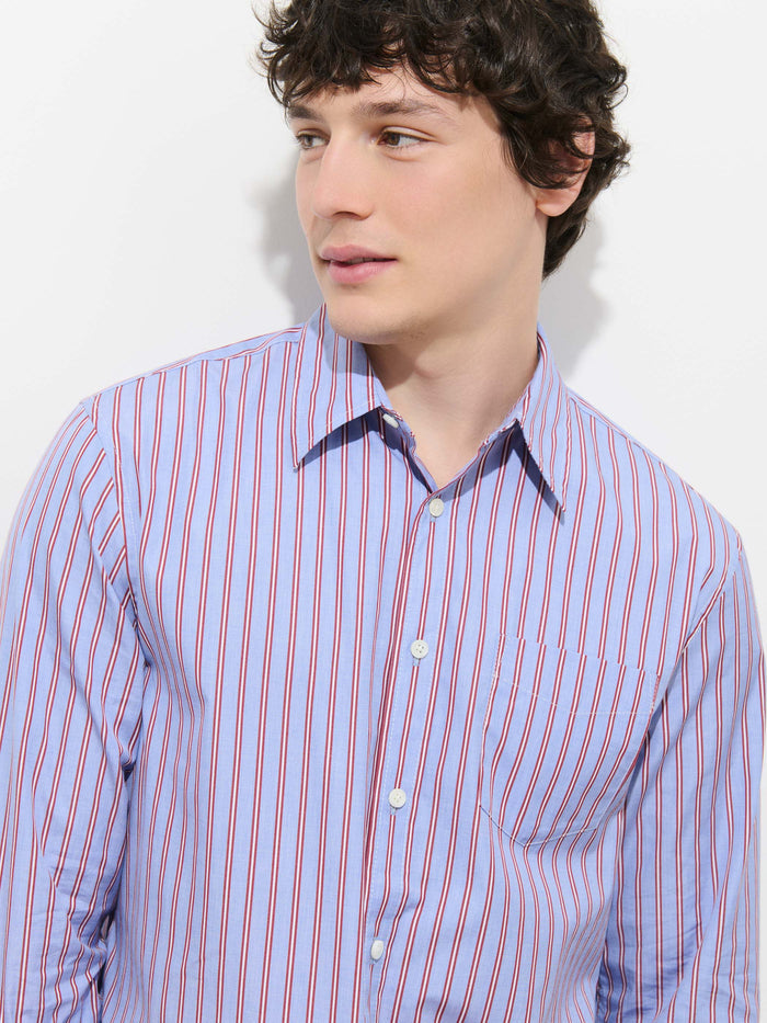 A young man with wavy brown hair looks to his left, wearing the Mill Point Collar Striped Shirt in Cotton Poplin—a blue shirt featuring red and white vertical stripes, crafted from 100% cotton—while standing against a plain white background.