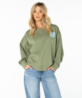 A woman stands with her hands in her pockets facing forward against a plain white background, wearing the Roxy Morning Stroll Top in Oil Green Wave Riders Club and light blue jeans.