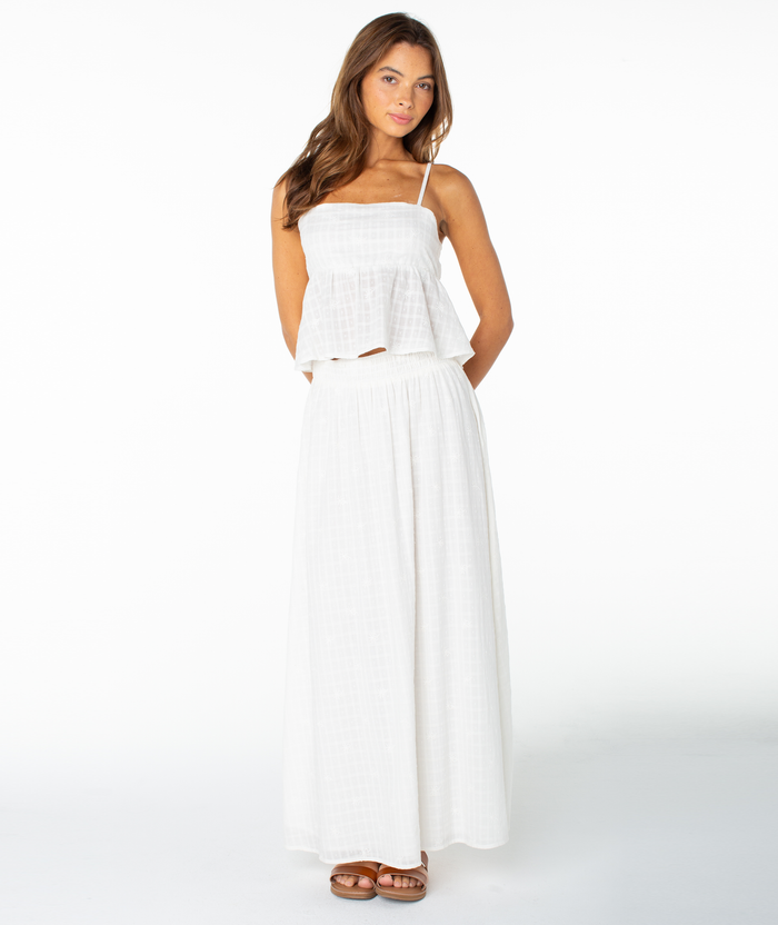A woman wearing the Roxy Pierside Top—a flattering, white, sleeveless spaghetti strap top—paired with a long, flowy white skirt stands against a plain white background, looking slightly to the side with her hands behind her back.