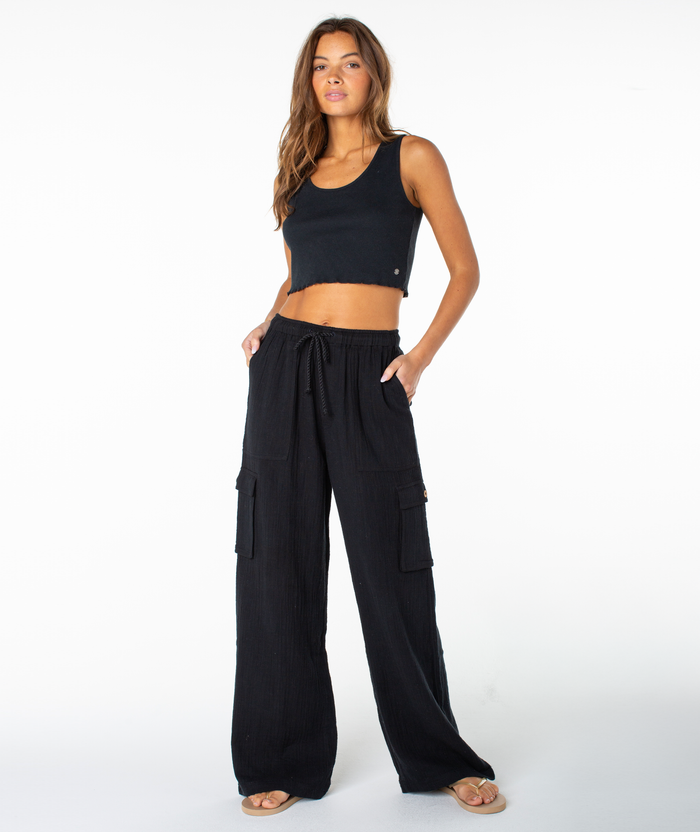 A woman stands against a white background wearing a black crop top, sandals, and the Roxy Precious Cargo Slub Pant—a pair of black wide-leg cargo pants with pockets. With her hands in her pockets and looking slightly to the side, she effortlessly showcases these breathable pants for a comfortable, modern look.