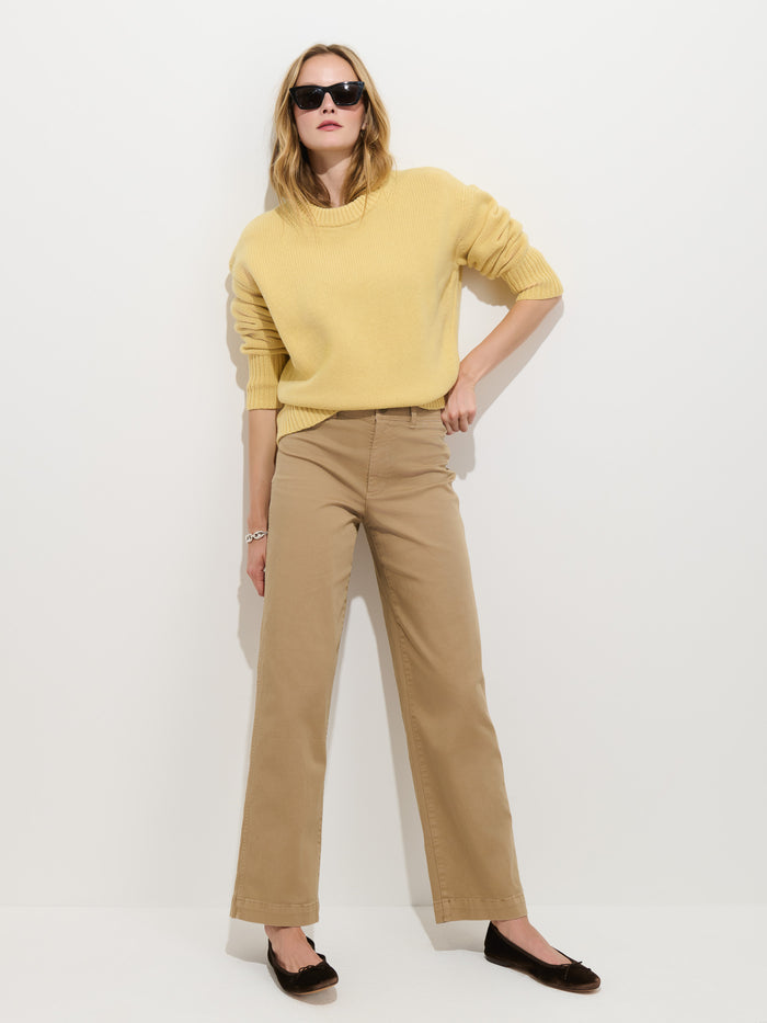 A person wearing sunglasses, a yellow sweater, the Sutton Pant in Vintage Twill, and dark flats stands against a plain white wall with hands in pockets.