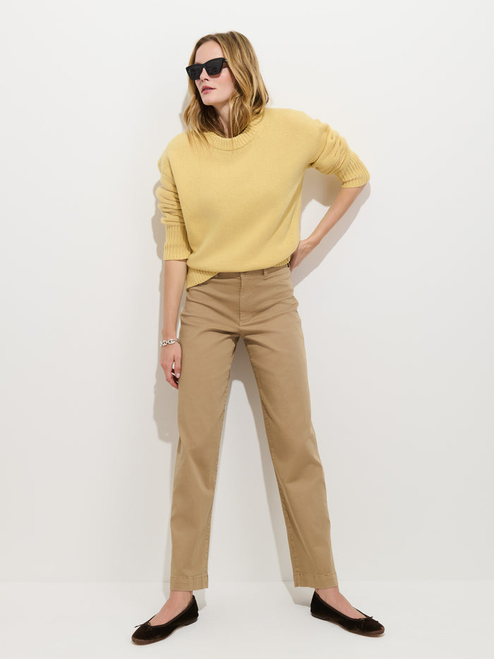 A woman in sunglasses stands against a white wall with her hands on her hips and feet apart, wearing a yellow sweater, black flats, and the Sutton Pant in Vintage Twill.