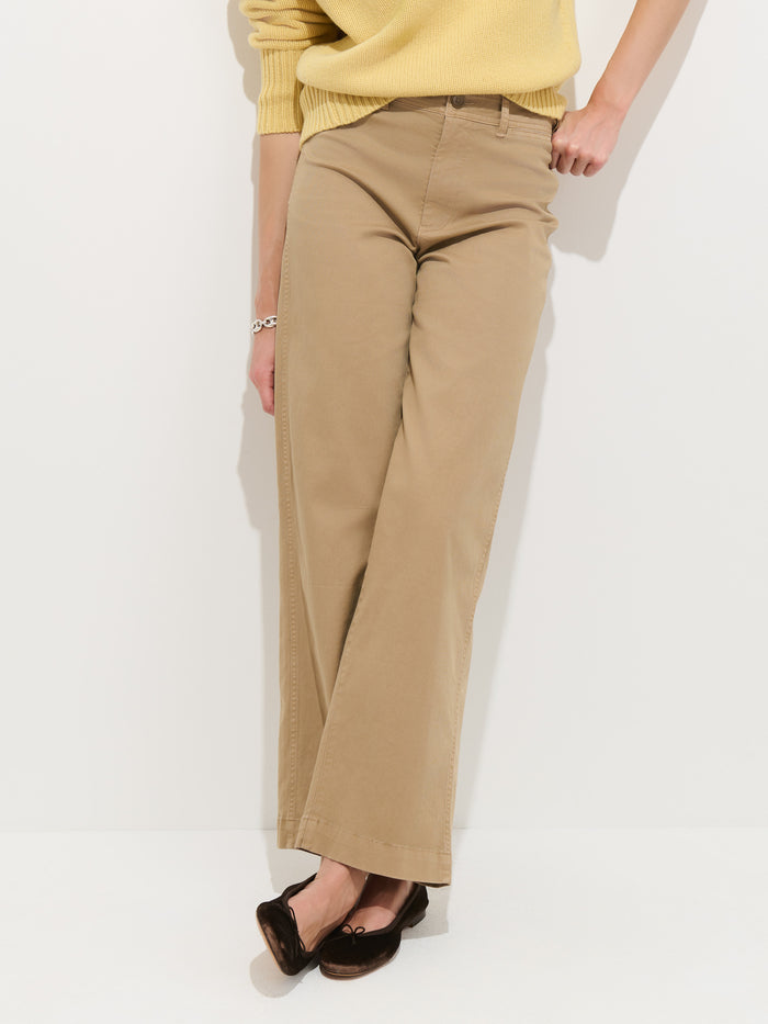 A person wears the Sutton Pant in Vintage Twill—tan high-rise, wide-leg chinos—with a yellow sweater and dark loafers, standing against a white background with one leg crossed over the other. The upper body is partially out of frame.
