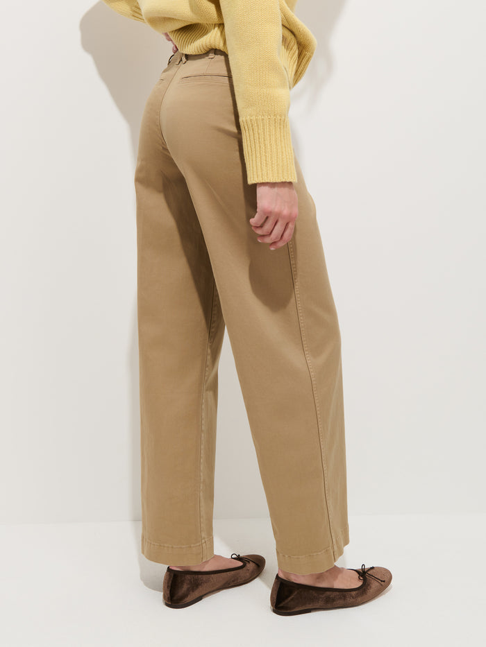 A person stands in profile against a plain white background, one arm relaxed, wearing the Sutton Pant in Vintage Twill—a tan high-rise wide-leg chino—paired with a yellow sweater and brown ballet flats.