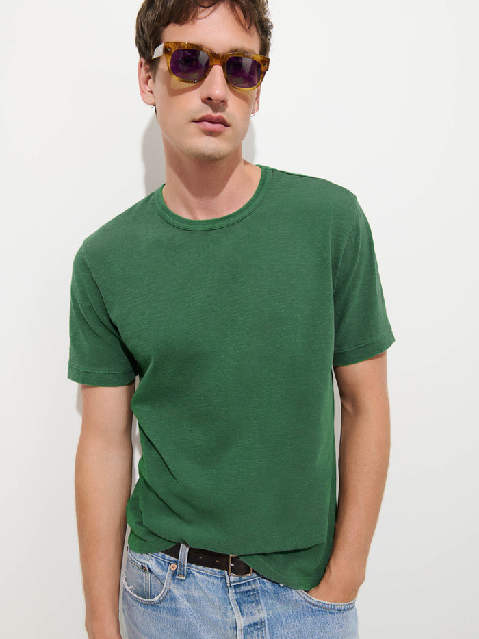 A man wearing sunglasses, The Standard T-Shirt In Slub Cotton—a green short-sleeve men’s tee made from breathable fabric—and light blue jeans stands against a plain white background.