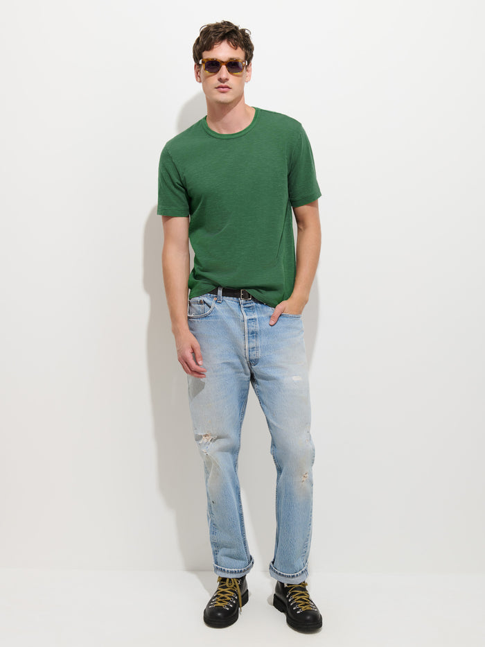 A man wearing sunglasses, The Standard T-Shirt In Slub Cotton in green, light blue ripped jeans, and black boots stands against a plain white background with one hand in his pocket.